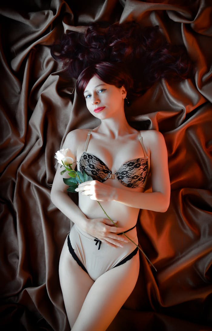 portfolio-img-04 A serene woman in lingerie lies on silk sheets, holding a white rose, exuding elegance and grace.